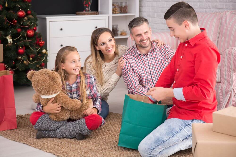otec, mama a sestra pozorujú chlapca ako rozbaľuje darček